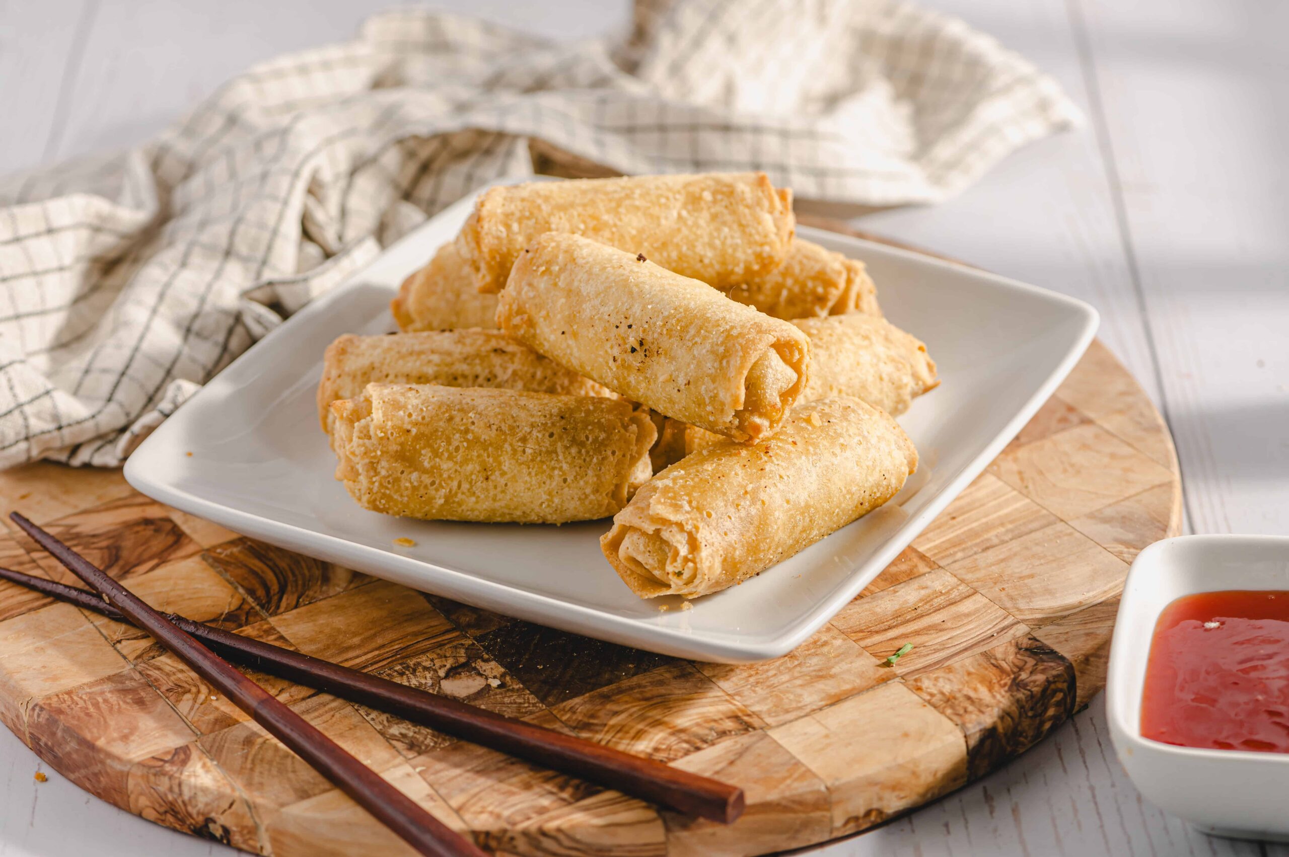 Crispy spring rolls with golden, crunchy exterior served on a white platter with a side of sweet chili dipping sauce, garnished with chopsticks on a rustic wooden board.