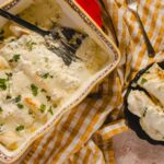Creamy chicken casserole with herbs in a colorful baking dish, topped with melted cheese, served in a black bowl with a spoon, surrounded by a checkered cloth and utensils for a delicious homemade dinner.