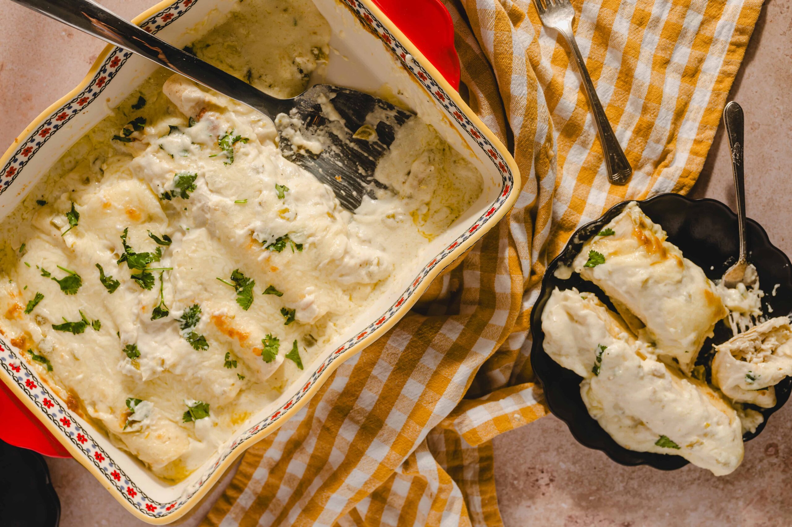 Creamy chicken casserole with herbs in a colorful baking dish, topped with melted cheese, served in a black bowl with a spoon, surrounded by a checkered cloth and utensils for a delicious homemade dinner.