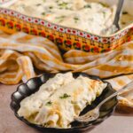 Creamy chicken and cheese enchiladas served in a black dish, topped with melted cheese and garnished with parsley, with an authentic Mexican casserole dish of enchiladas in the background.