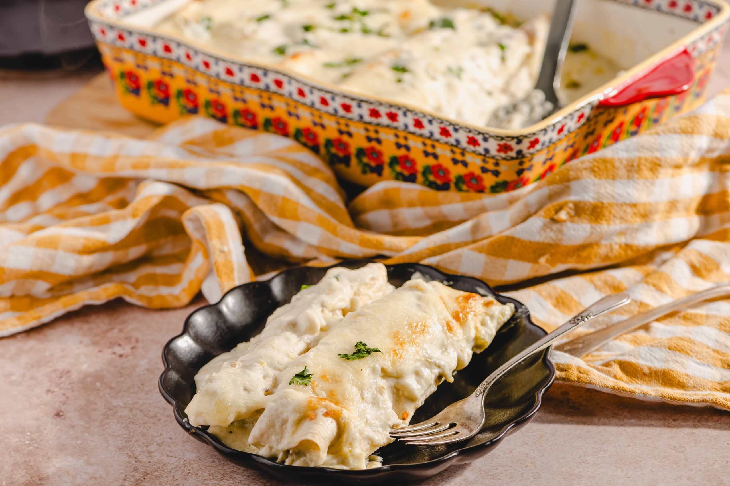 Creamy chicken and cheese enchiladas served in a black dish, topped with melted cheese and garnished with parsley, with an authentic Mexican casserole dish of enchiladas in the background.