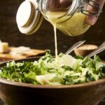 Creamy dressing being poured over a fresh green salad in a rustic wooden bowl, with cheese and kitchen tools in the background, perfect for healthy recipes.