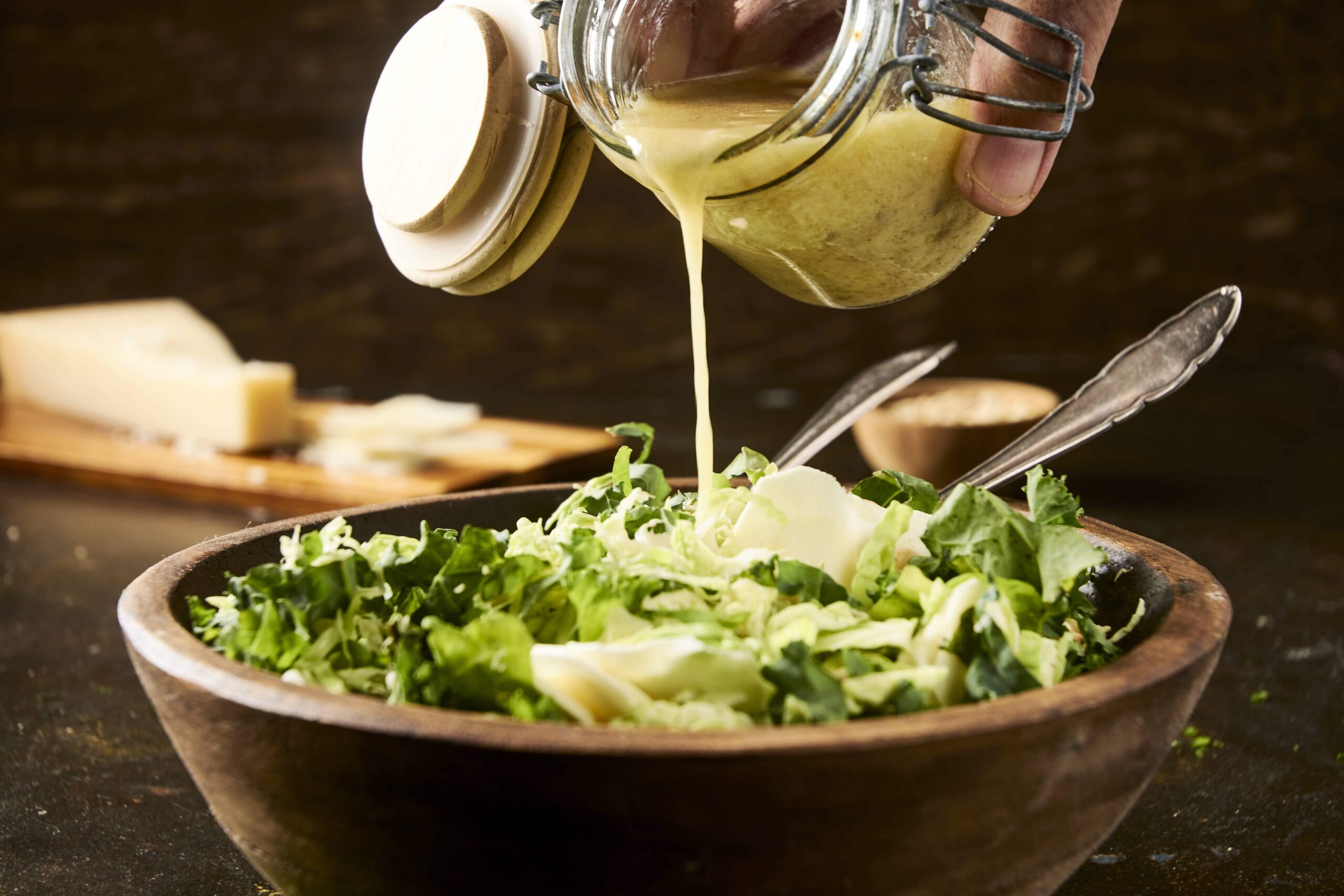 Creamy dressing being poured over a fresh green salad in a rustic wooden bowl, with cheese and kitchen tools in the background, perfect for healthy recipes.