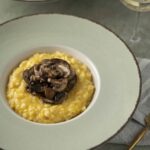 Creamy mushroom risotto topped with sautéed mushrooms, served in a stylish bowl on a grey table with a gold fork and a glass of white wine. Perfect dish for mushroom lovers and comfort food enthusiasts.