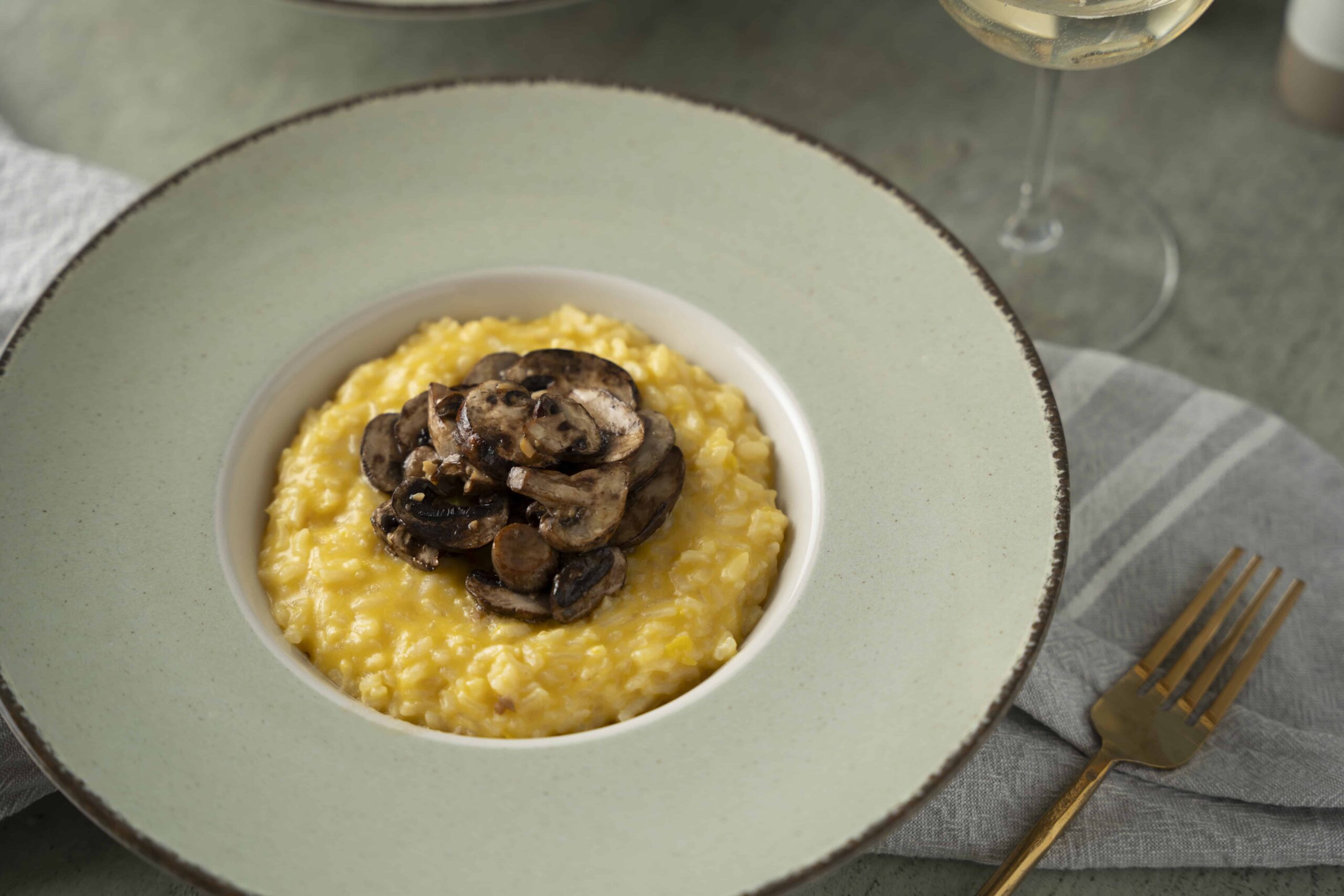 Creamy mushroom risotto topped with sautéed mushrooms, served in a stylish bowl on a grey table with a gold fork and a glass of white wine. Perfect dish for mushroom lovers and comfort food enthusiasts.