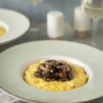 Creamy mushroom risotto served on a stylish green-rimmed plate with sliced mushrooms on top, accompanied by a glass of white wine and salt and pepper shakers in the background, perfect for a gourmet dinner.