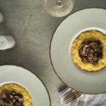 Creamy mushroom risotto served in a bowl with sautéed mushrooms, plated on a stylish salad plate, accompanied by wine glasses and salt and pepper shakers on a rustic table.