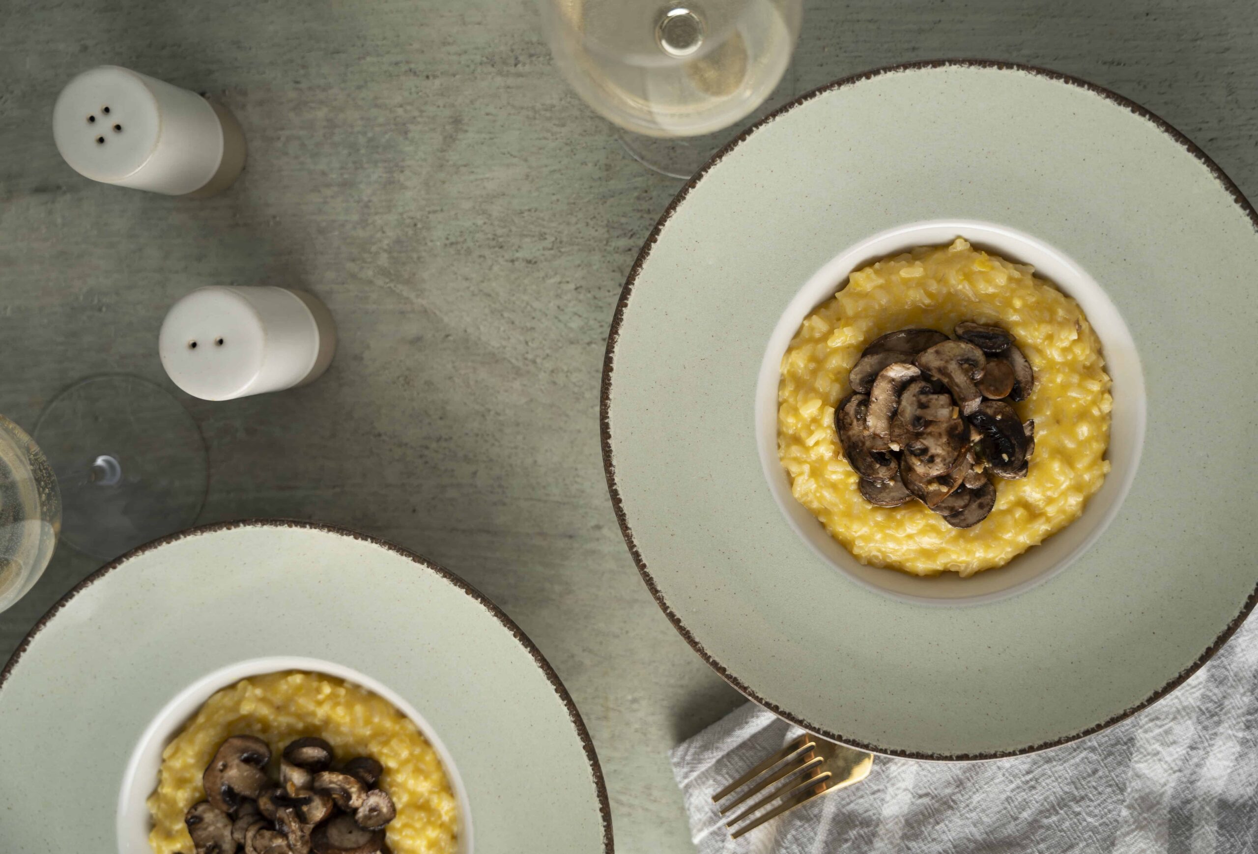 Creamy mushroom risotto served in a bowl with sautéed mushrooms, plated on a stylish salad plate, accompanied by wine glasses and salt and pepper shakers on a rustic table.