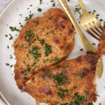 Pan-fried breaded chicken breast with chopped parsley on a white ceramic plate, golden crispy crust, served with fork and knife, close-up of delicious homemade chicken dish, perfect for healthy dinner.