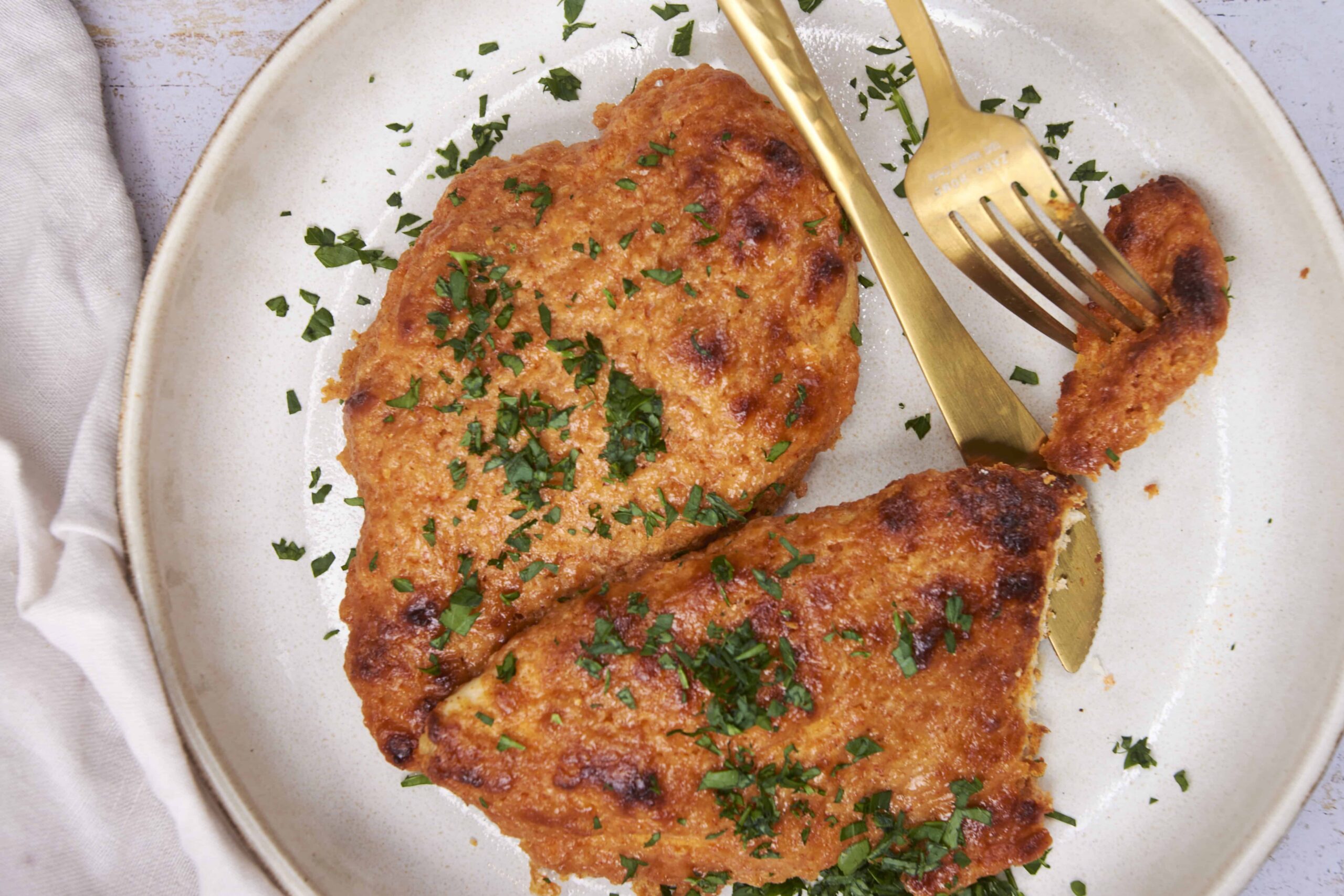 Pan-fried breaded chicken breast with chopped parsley on a white ceramic plate, golden crispy crust, served with fork and knife, close-up of delicious homemade chicken dish, perfect for healthy dinner.