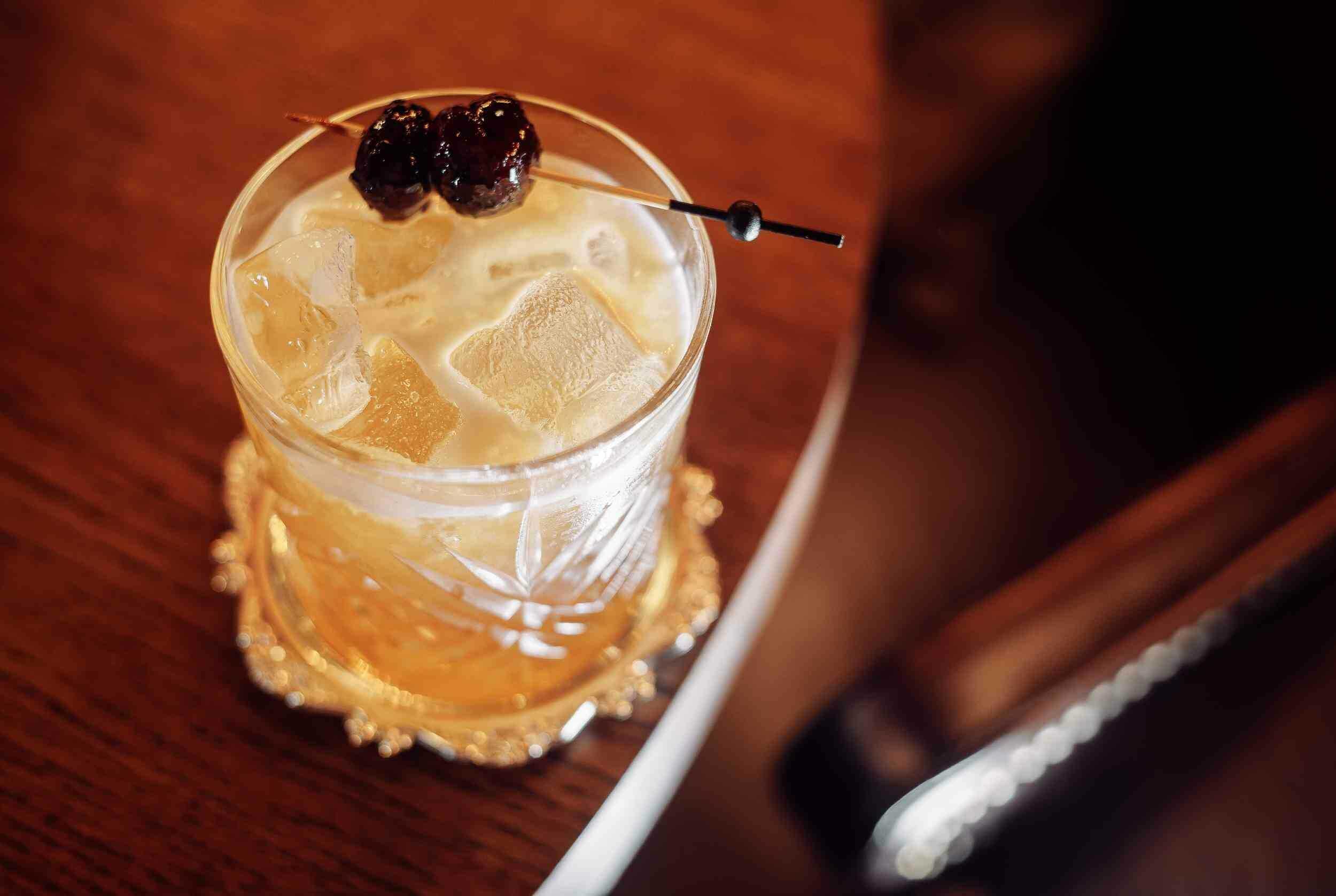 Refreshing whiskey cocktail with ice cubes and dark cherries on a wooden table, served in an elegant glass.