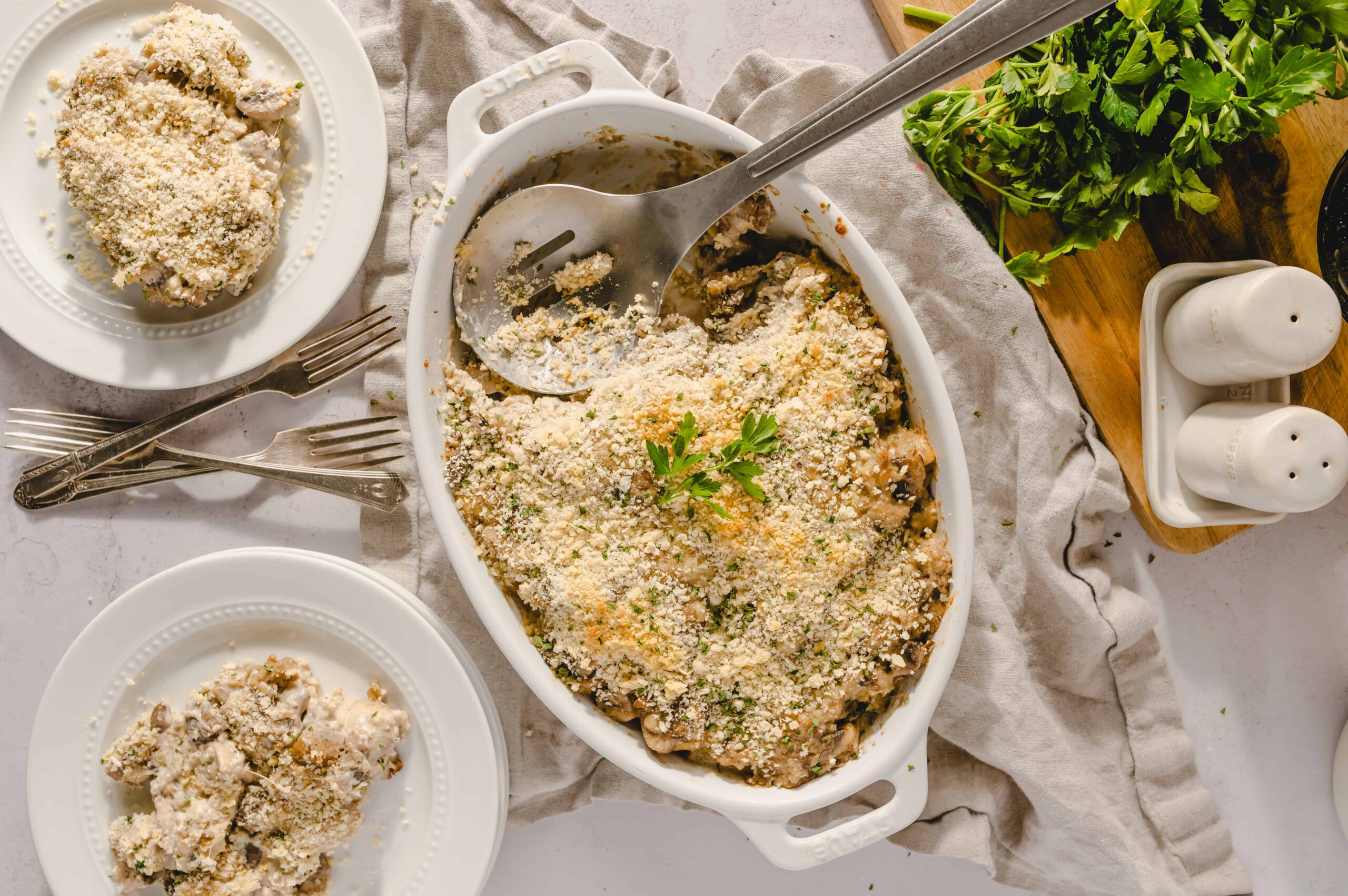 Creamy chicken casserole with breadcrumbs and herbs, served in a white dish with fresh parsley garnish, accompanied by plates of chicken, perfect for comfort food recipes.