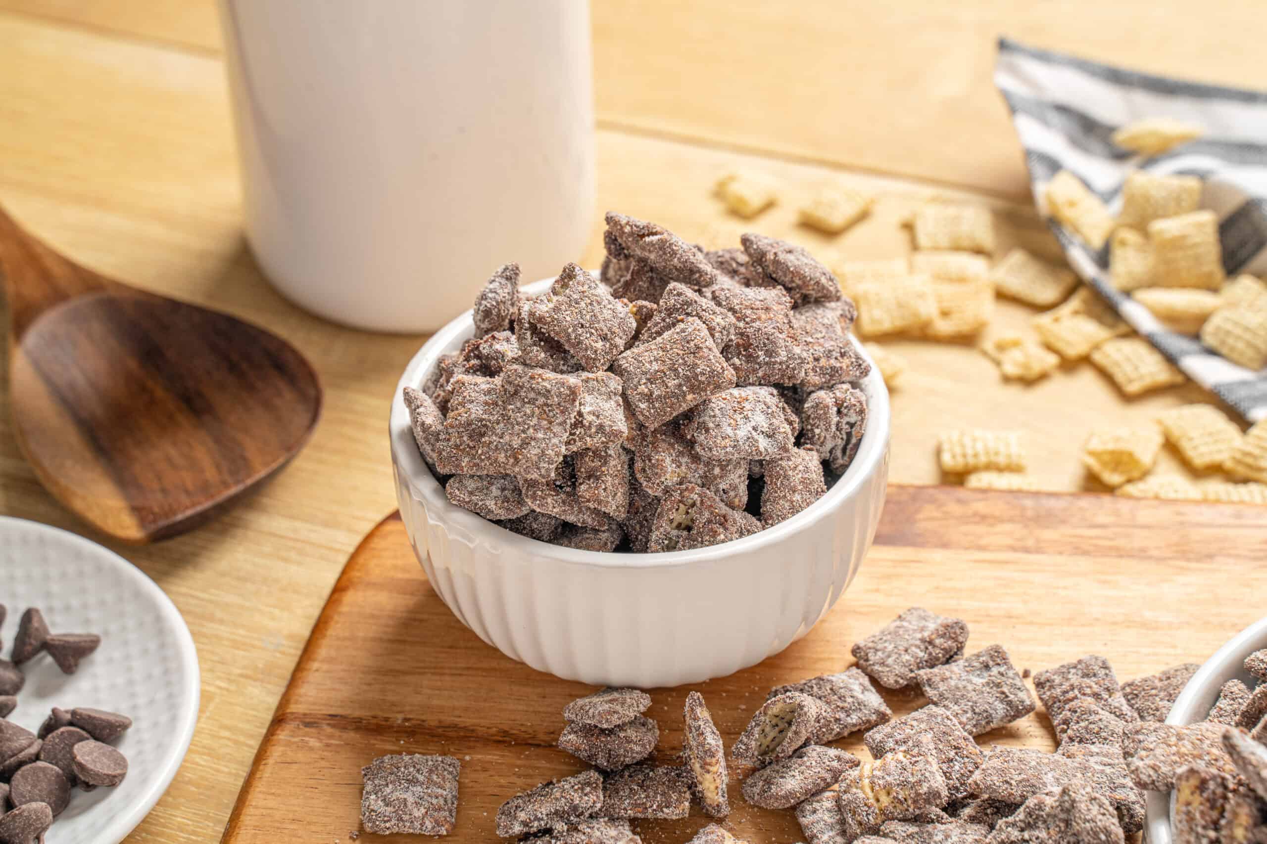 Sweet chocolate-covered cereal squares in a white bowl with scattered cereal pieces and a glass of milk on a wooden table. Perfect for breakfast or snacks.