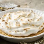 Fluffy lemon meringue pie topped with toasted meringue in a white ceramic dish, on a rustic wooden table with gold spoons and a dessert plate in the background.