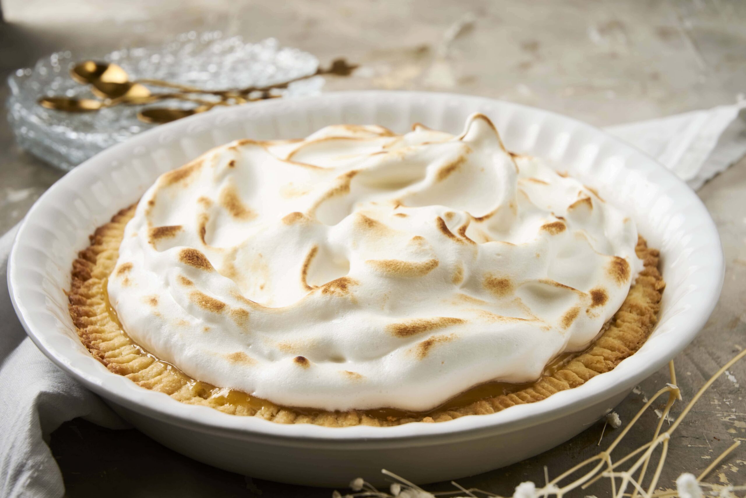 Fluffy lemon meringue pie topped with toasted meringue in a white ceramic dish, on a rustic wooden table with gold spoons and a dessert plate in the background.