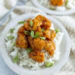 Crispy orange chicken over white rice with green onions and black sesame seeds, served on a rustic white plate, featuring Asian-inspired flavors and home-cooked meal presentation.