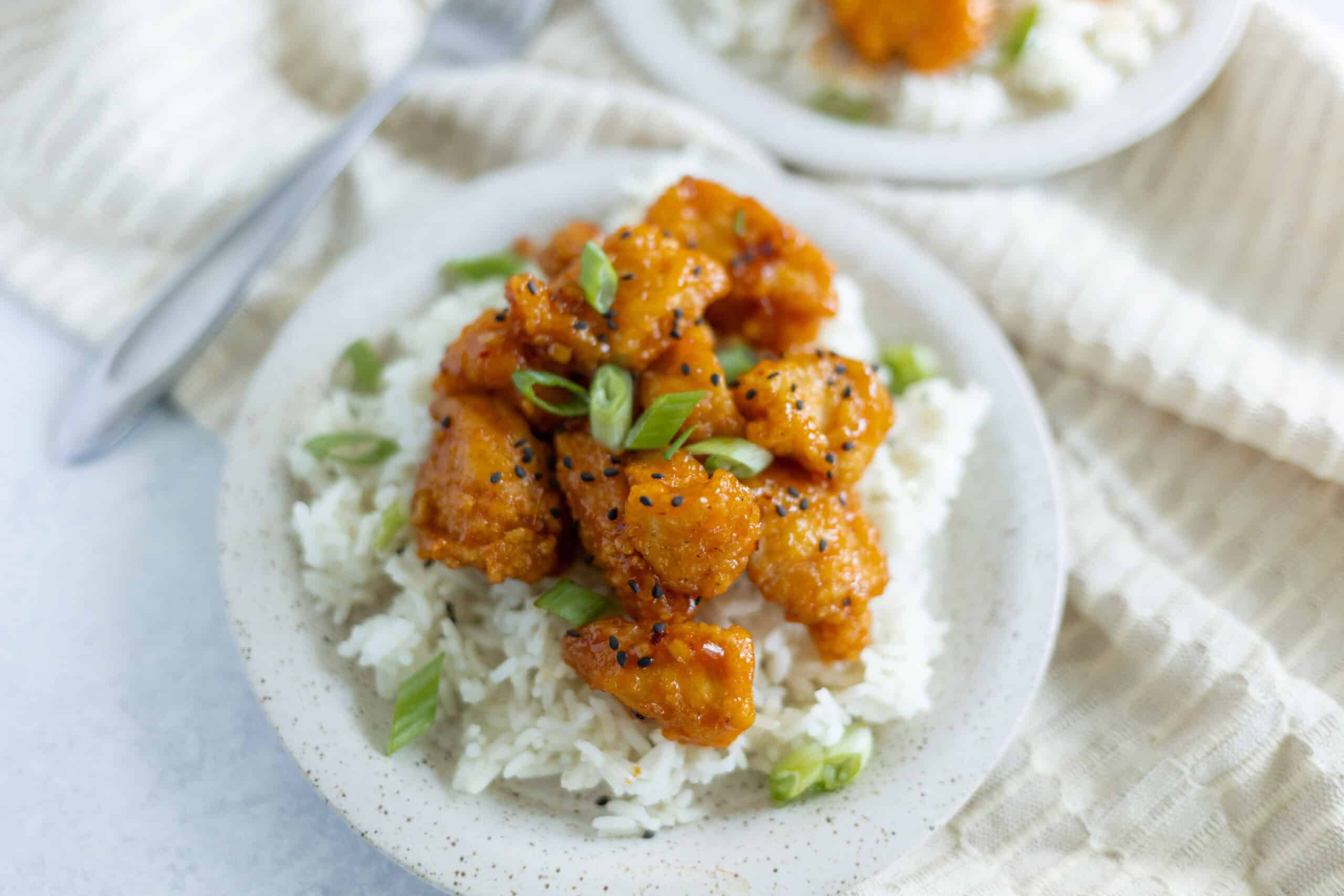 Crispy orange chicken over white rice with green onions and black sesame seeds, served on a rustic white plate, featuring Asian-inspired flavors and home-cooked meal presentation.