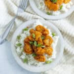 Spicy orange chicken served over white rice garnished with green onions and black sesame seeds on a speckled plate.