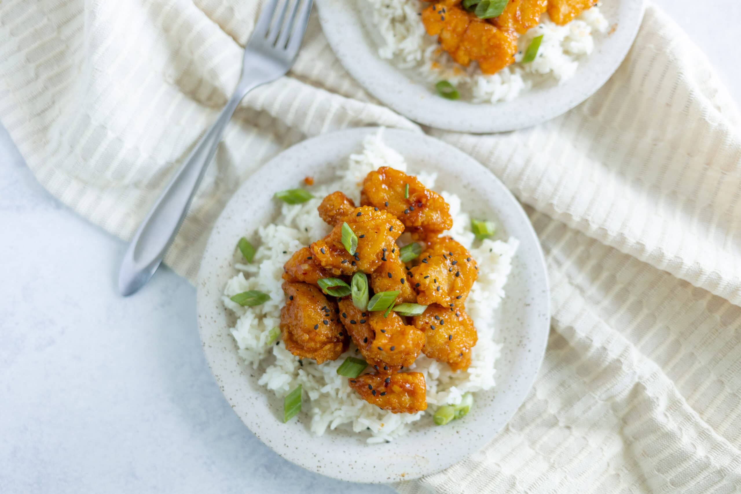 Spicy orange chicken served over white rice garnished with green onions and black sesame seeds on a speckled plate.