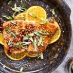 Salmon fillet with orange slices, sesame seeds, and green onions on a dark rustic plate, garnished with fresh orange wedges; healthy seafood recipe.