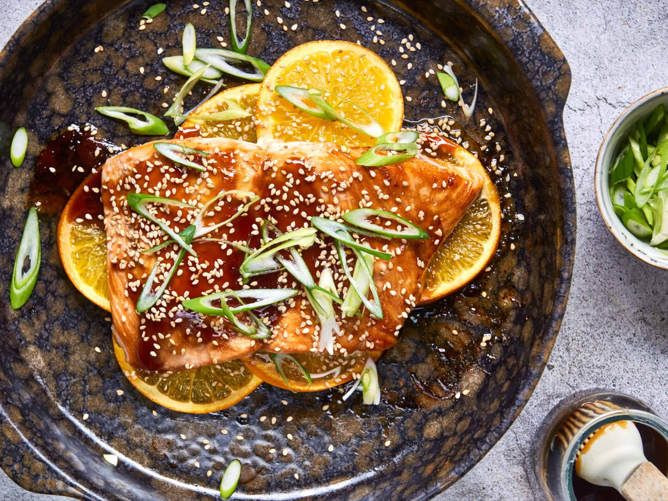 Salmon fillet with orange slices, sesame seeds, and green onions on a dark rustic plate, garnished with fresh orange wedges; healthy seafood recipe.