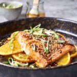 Baked salmon fillet topped with sesame seeds, drizzled with sauce, served on a bed of orange slices topped with green onions in a rustic black skillet.