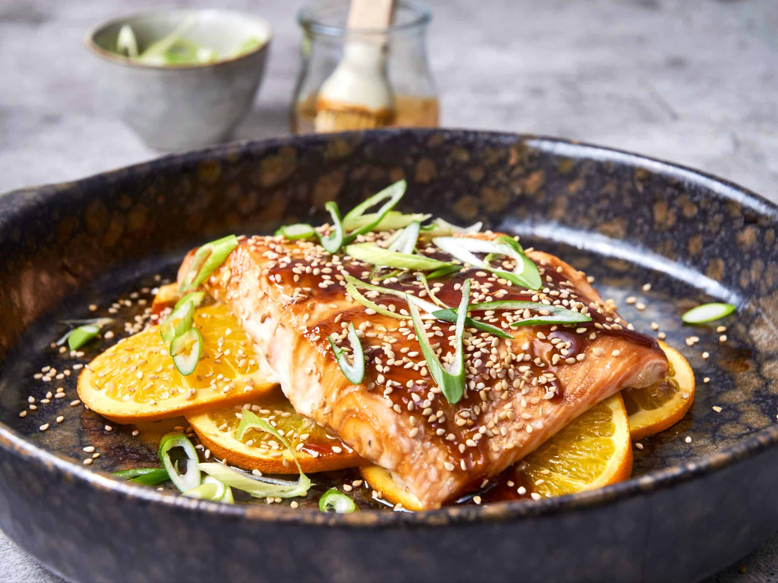 Baked salmon fillet topped with sesame seeds, drizzled with sauce, served on a bed of orange slices topped with green onions in a rustic black skillet.