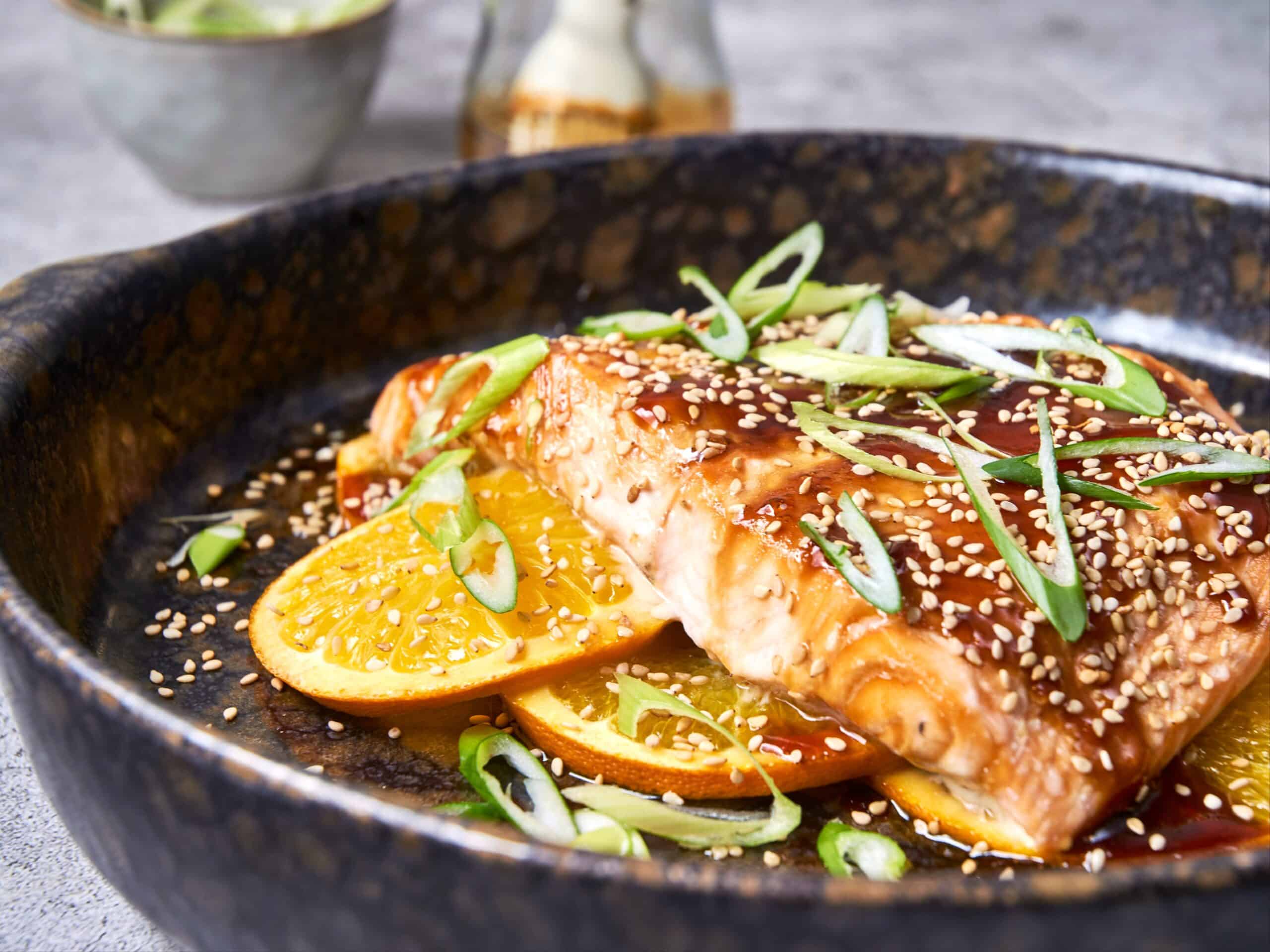 Sushi with fresh salmon, orange slices, green onions, sesame seeds, and sauce served in a rustic black bowl, highlighting healthy, flavorful seafood dishes.