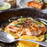 Fresh salmon fillet with citrus slices, garnished with green onions and sesame seeds, served in a black skillet with savory sauce, perfect for healthy, flavorful seafood recipes.