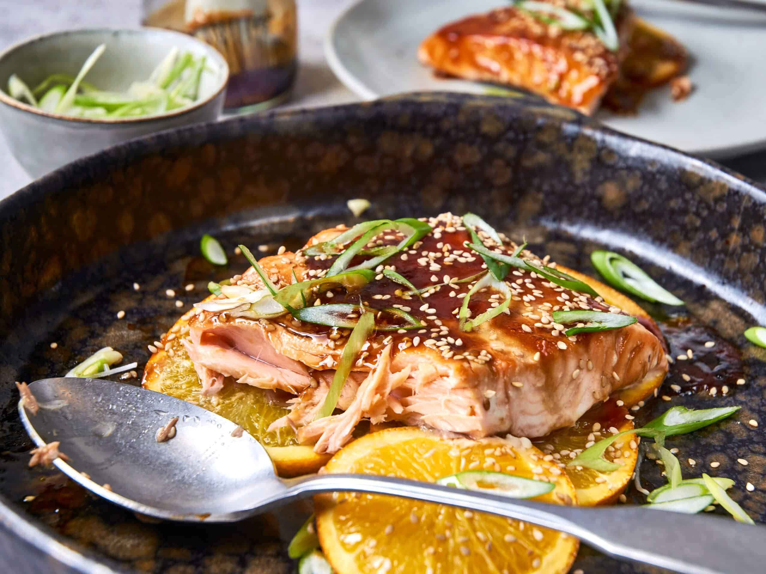 Fresh salmon fillet with citrus slices, garnished with green onions and sesame seeds, served in a black skillet with savory sauce, perfect for healthy, flavorful seafood recipes.
