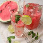 Refreshing watermelon lemonade with fresh mint and lime served in a glass, with a watermelon half and lime slices on a white wooden surface.