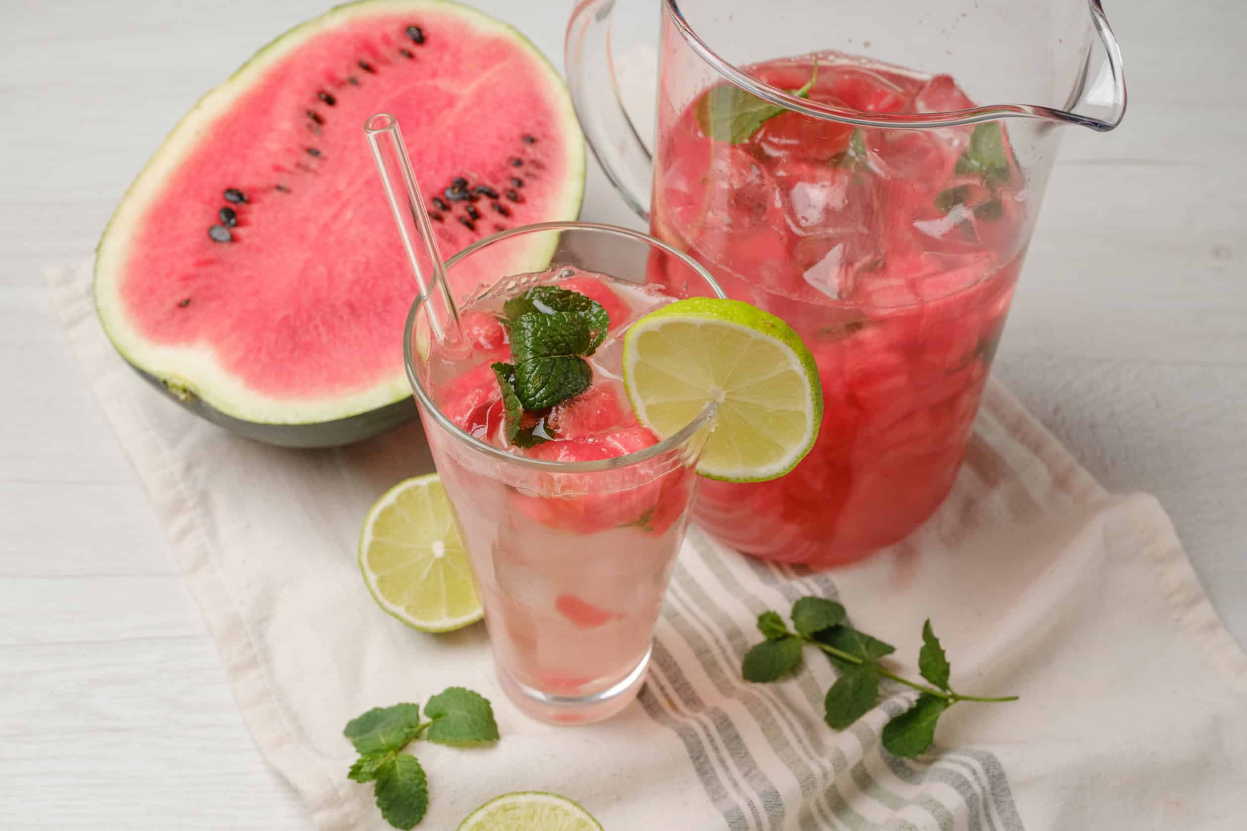 Refreshing watermelon lemonade with fresh mint and lime served in a glass, with a watermelon half and lime slices on a white wooden surface.