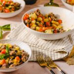 Vegetable and sausage potato salad in white bowls on a pink surface with gold forks and rustic table setting.