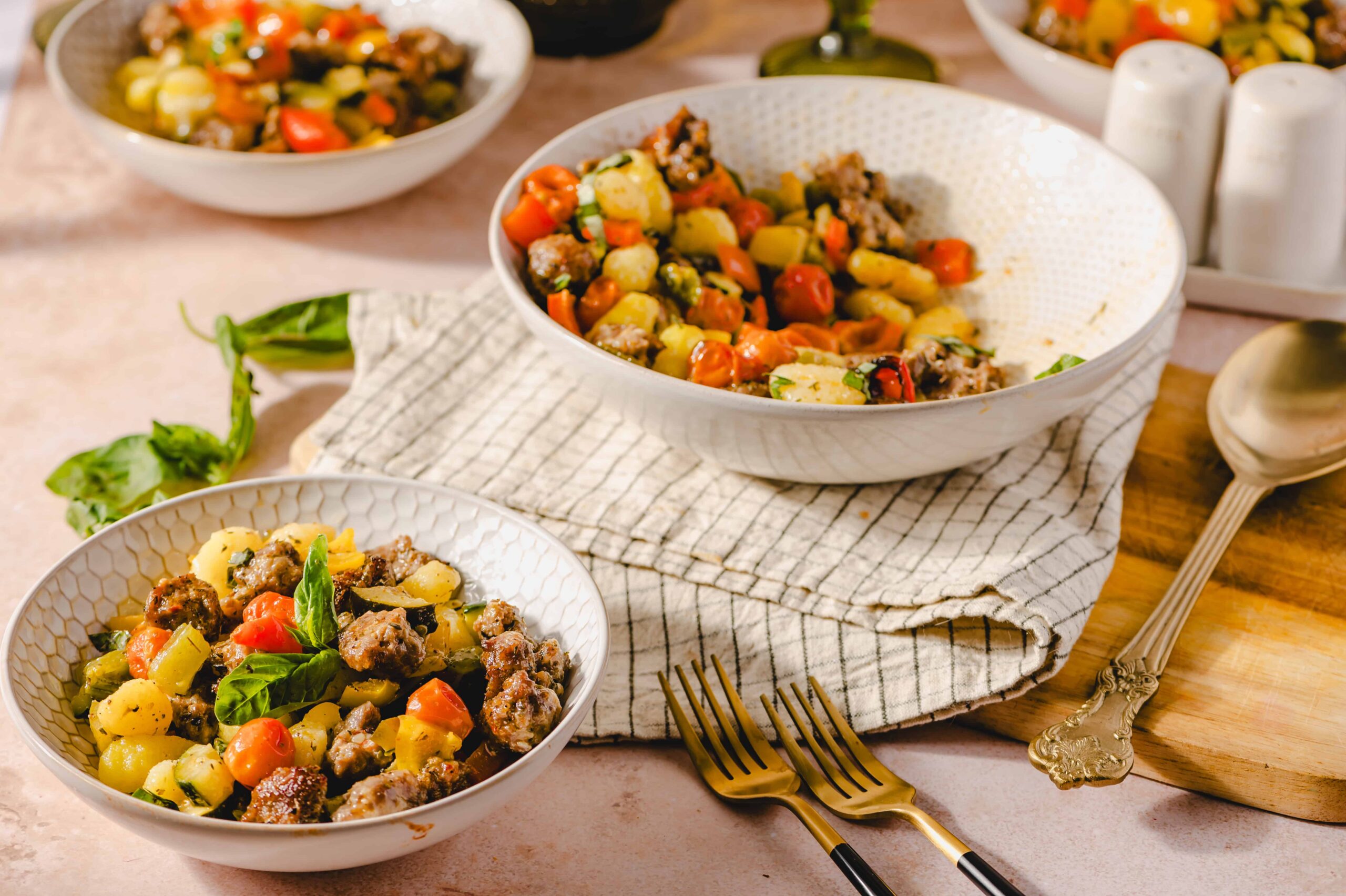 Vegetable and sausage potato salad in white bowls on a pink surface with gold forks and rustic table setting.