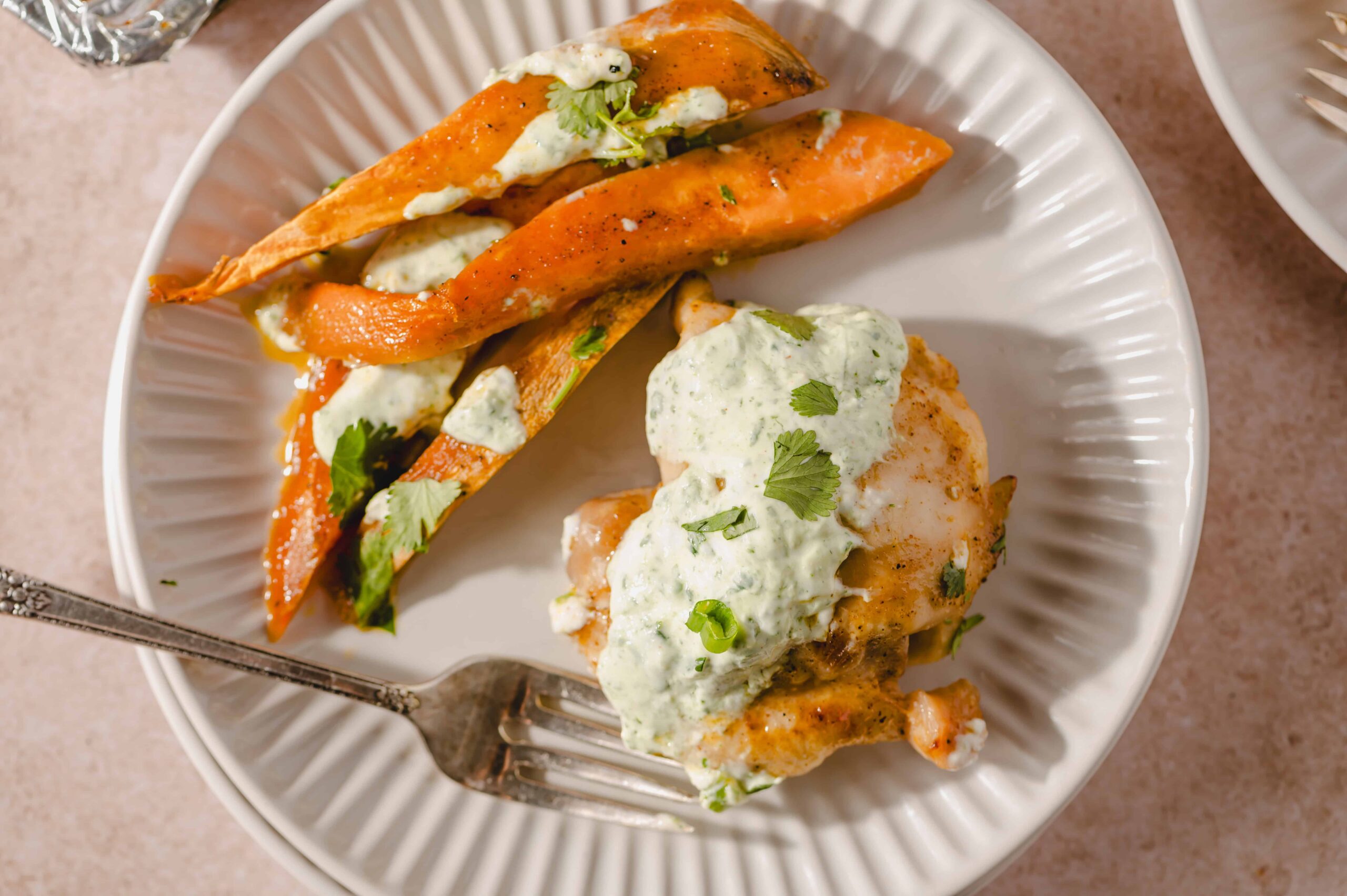 Roasted sweet potato wedges with cilantro lime sauce served alongside baked chicken topped with creamy herb dressing on a white plate.