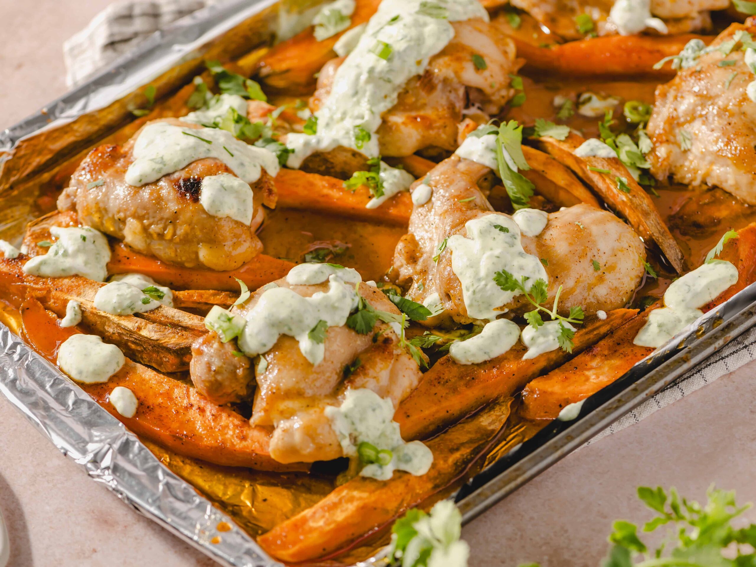 Roasted sweet potatoes with chicken, topped with cilantro and a creamy, herb-infused sauce, served on a baking sheet with aluminum foil for a delicious and healthy meal.