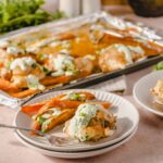 Roasted chicken with creamy herb sauce and roasted vegetables on a white plate. Baked chicken thighs served with a drizzle of herbed cream sauce, accompanied by roasted bell peppers, on a light pink surface.