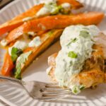 Golden-brown baked sweet potatoes topped with creamy herb sauce, served with fresh cilantro and green onions, on a white plate. Perfect for healthy, flavorful sweet potato recipes.