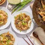Steamed chicken and vegetable stir-fry with noodles served on white plates, topped with chopped green onions, on a marble table with garlic, scallions, and wooden utensils.