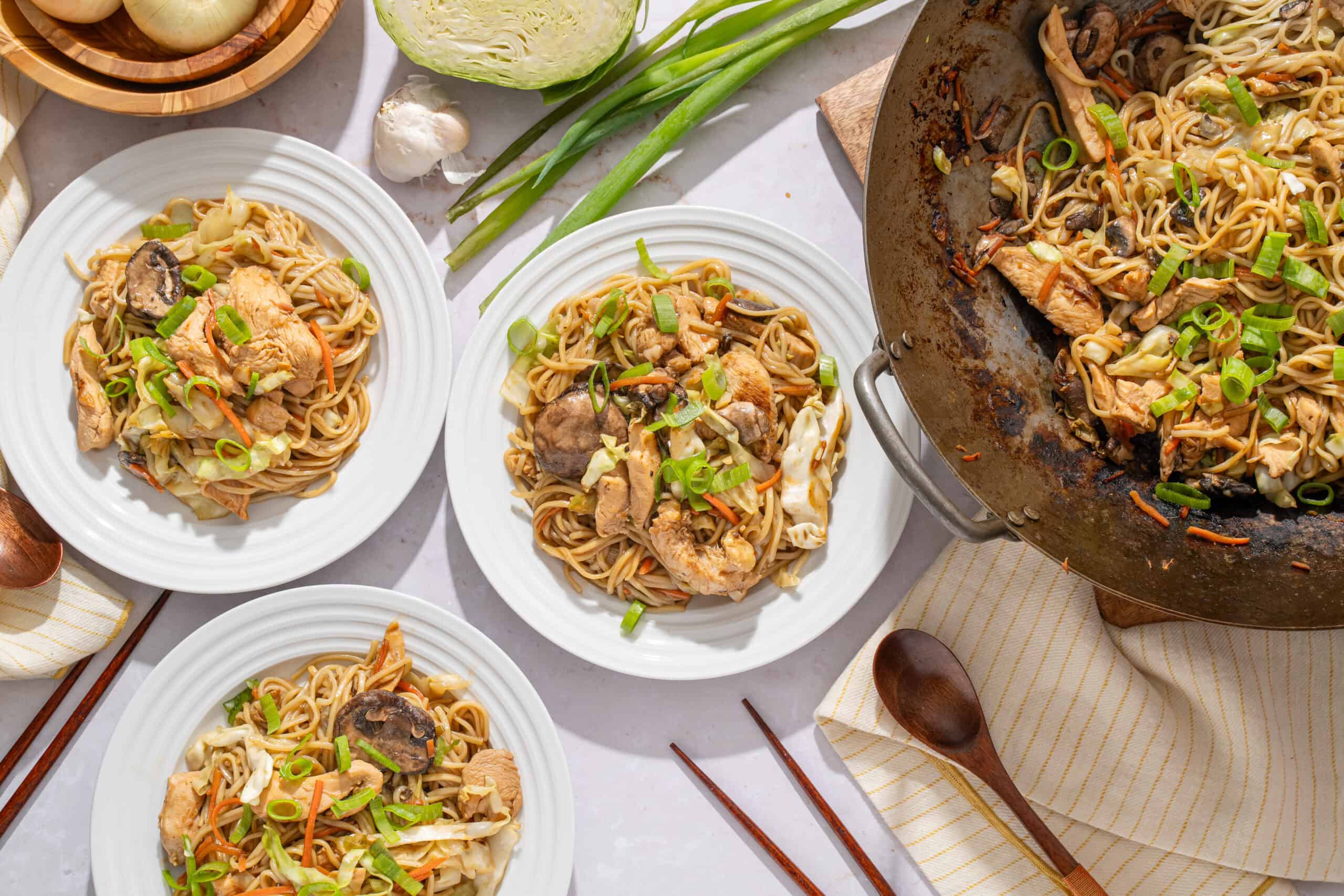 Steamed chicken and vegetable stir-fry with noodles served on white plates, topped with chopped green onions, on a marble table with garlic, scallions, and wooden utensils.