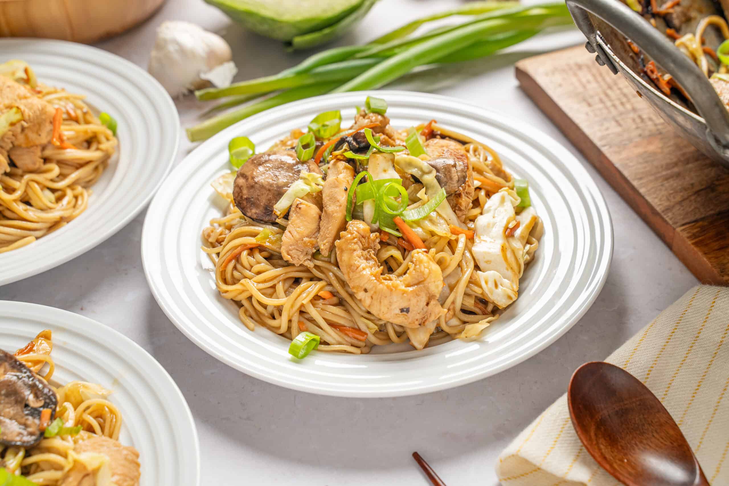 Savory chicken and vegetable stir-fry with noodles on white plate, garnished with green onions, served with assorted Asian-inspired dishes.