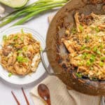 Stir-fried chicken and vegetable lo mein served on white plates with fresh green onions and garlic, featuring a large wok with noodles and ingredients in the background.