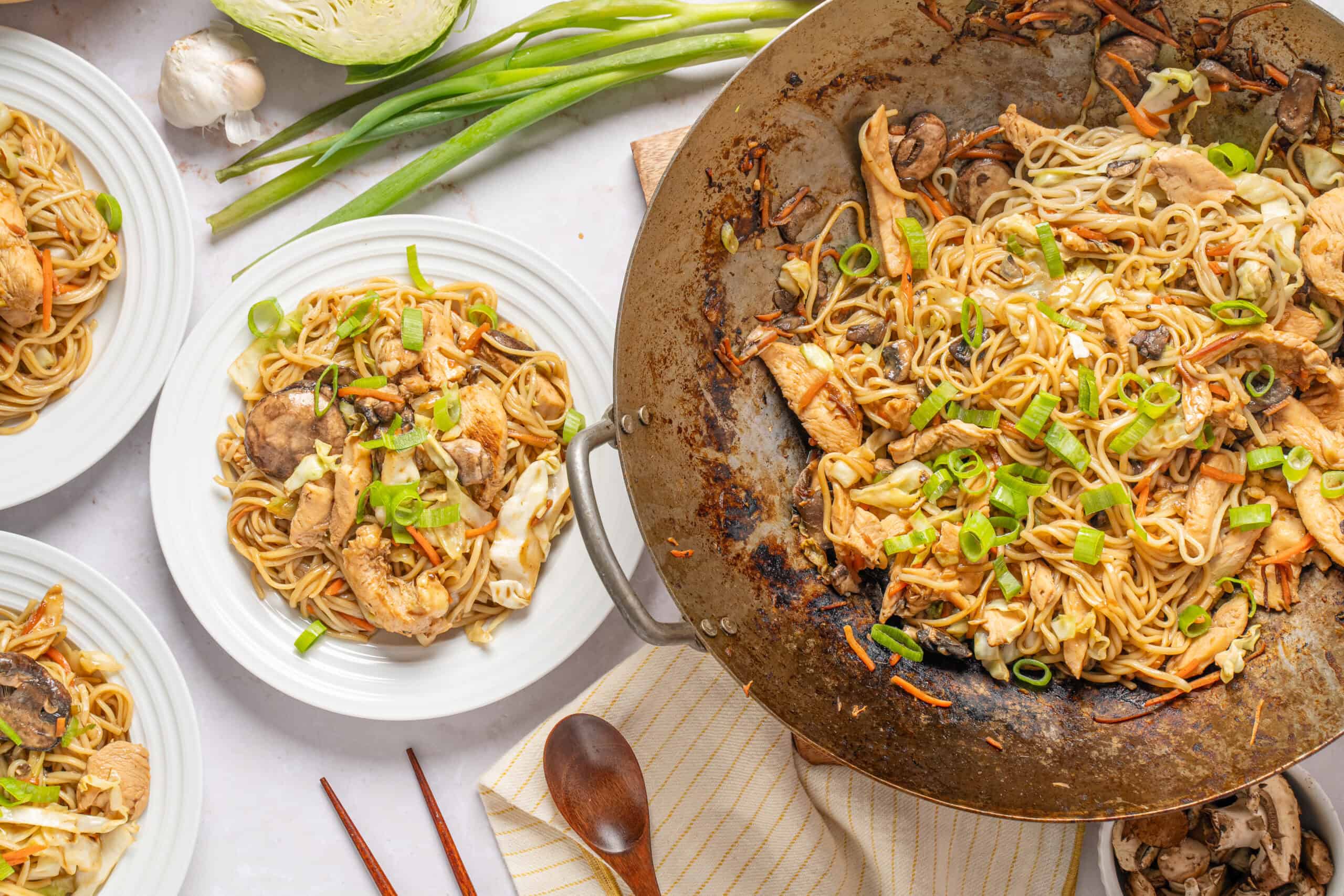 Stir-fried chicken and vegetable lo mein served on white plates with fresh green onions and garlic, featuring a large wok with noodles and ingredients in the background.