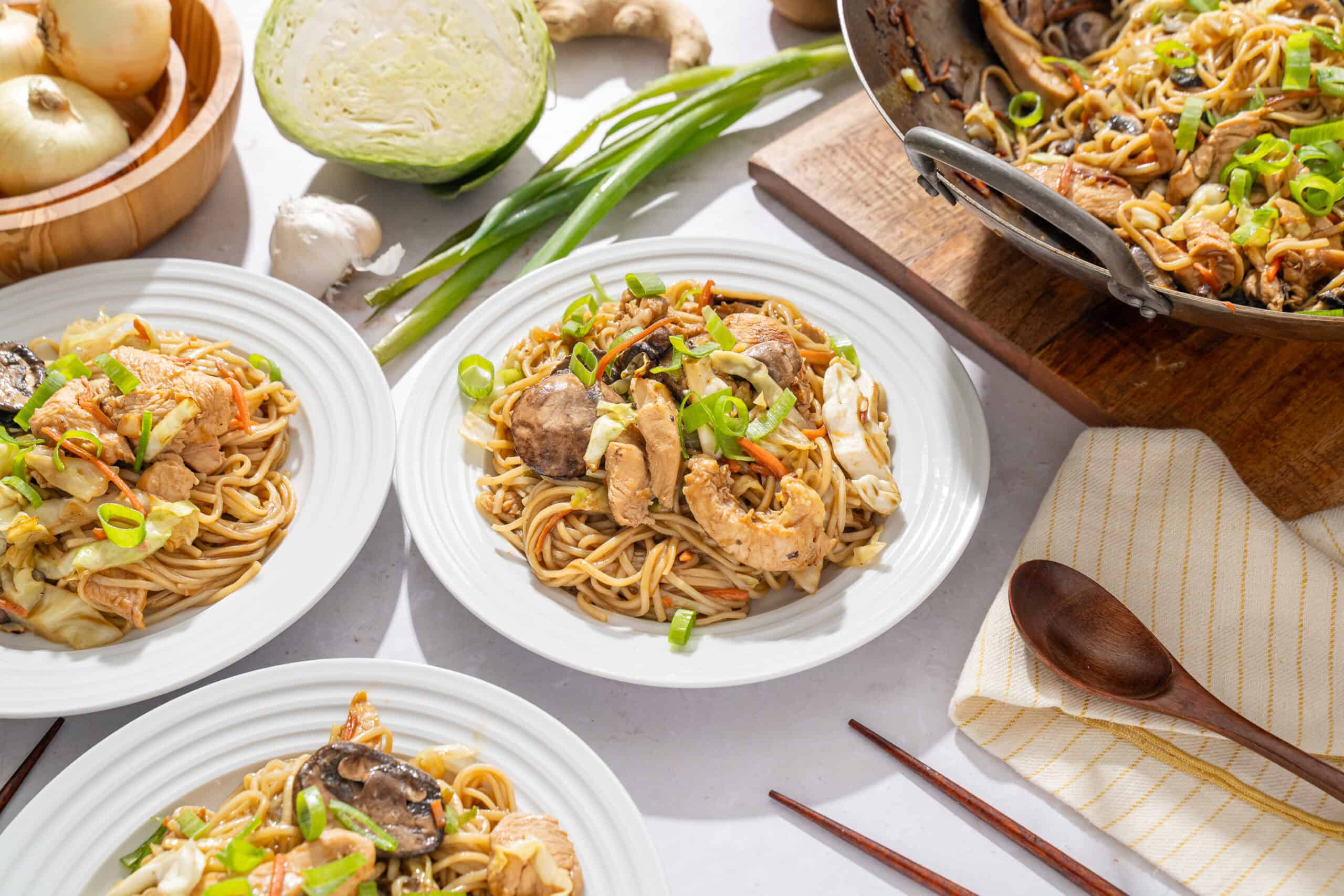 Stir-fried Asian noodle dishes with chicken, vegetables, and green onions served on white plates, showcasing a flavorful and colorful presentation.
