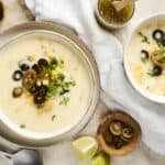 Creamy white cheesy soup topped with black olives, jalapeños, corn, and fresh cilantro, served in white bowls with lime wedges and rustic background.