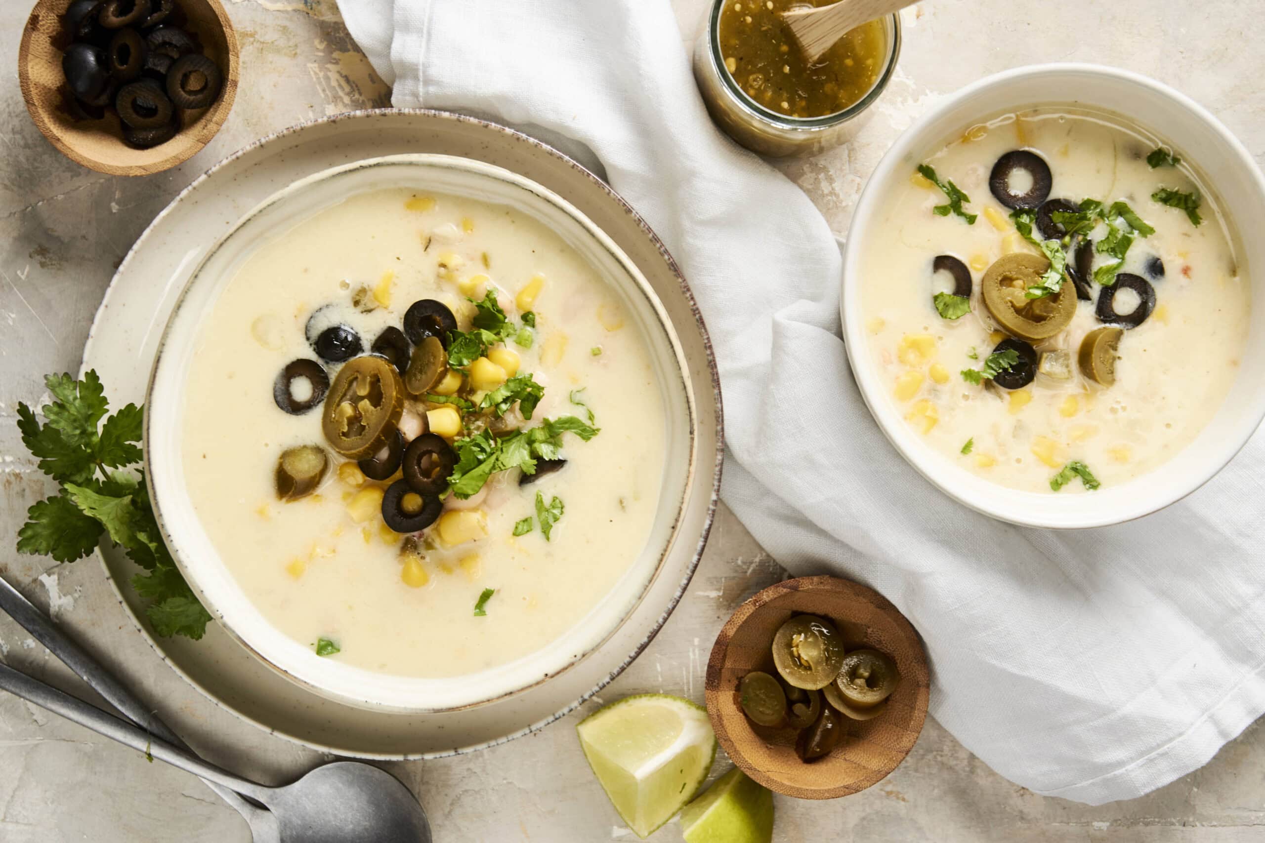 Creamy white cheesy soup topped with black olives, jalapeños, corn, and fresh cilantro, served in white bowls with lime wedges and rustic background.