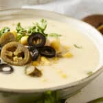 Creamy corn and black olive soup garnished with fresh cilantro and lemon wedges, served in a white bowl with a rustic backdrop, showcasing delicious Mexican-inspired flavors.