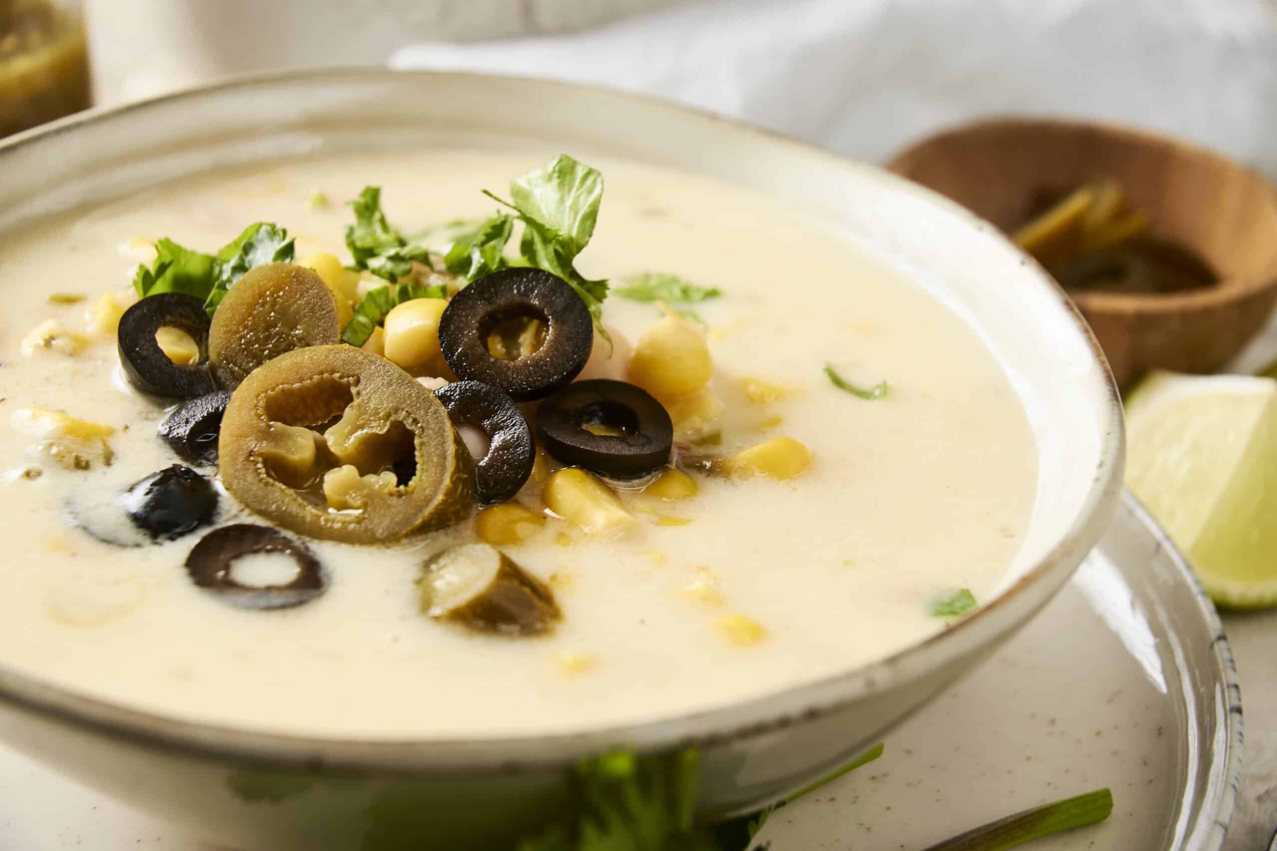 Creamy corn and black olive soup garnished with fresh cilantro and lemon wedges, served in a white bowl with a rustic backdrop, showcasing delicious Mexican-inspired flavors.
