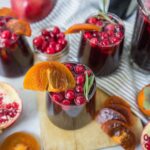 Fresh pomegranate and cranberry juice with slices of orange and garnishing herbs in tall glasses on a rustic wooden board and striped cloth, featuring vibrant seasonal fruits and beverage presentation.
