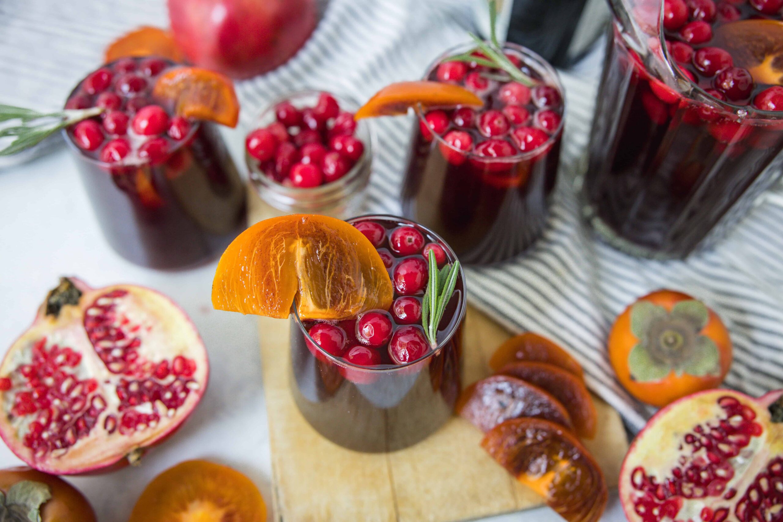 Fresh pomegranate and cranberry juice with slices of orange and garnishing herbs in tall glasses on a rustic wooden board and striped cloth, featuring vibrant seasonal fruits and beverage presentation.